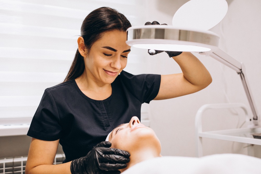 Dermatologista realiza avaliação facial em paciente deitada em maca, utilizando iluminação profissional em consultório dermatológico.